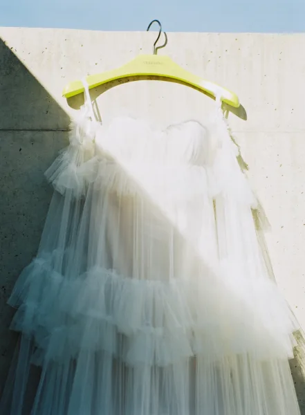 Wedding dress, tulle wedding dress hanging on a dress hanger with spaghetti straps and tiered ruffle skirt against a sunlit concrete wall