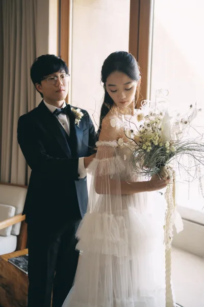 Couple portrait of bride holding bouquet as groom adjusts her off-the-shoulder tulle dress in soft window light by curtains and sofa