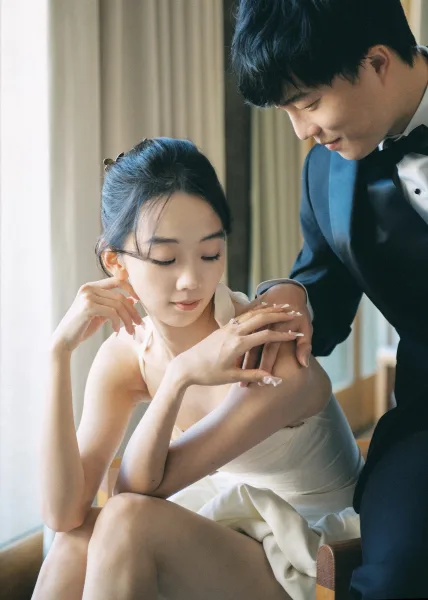 Couple portrait of groom kissing bride’s forehead as she sits in a strapless wedding dress, framed by sheer curtains and window light