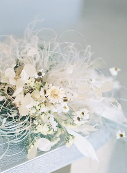Wedding bouquet with a daisy bridal bouquet look, featuring pampas grass accents and ribbon resting on a tabletop against a light blue wall