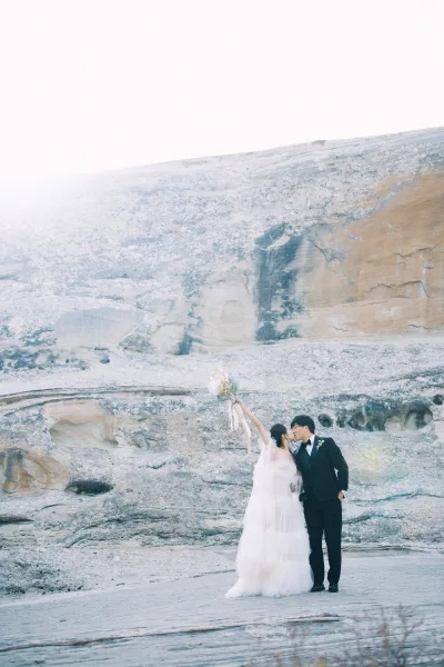Wedding kiss portrait of bride and groom kiss on a rocky cliffside, bride lifting bouquet with ribbon streamers, veil and tuxedo boutonniere under open sky
