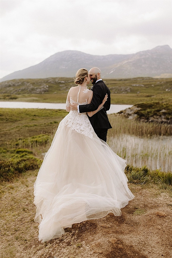 Couple portrait of bride and groom embracing, her lace tulle skirt and low bun visible, by a lakeside mountain field under cloudy sky