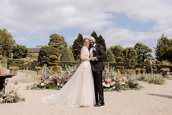 Couple portrait of bride and groom hugging in a formal garden, lace dress and long veil, groom in tuxedo with boutonniere, bouquet in hand