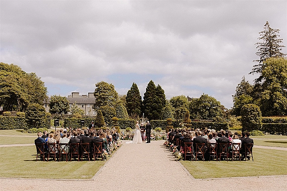 Outdoor wedding ceremony with wooden chairs in neat rows, aisle lined with flowers on a manicured lawn before garden hedges and an estate building
