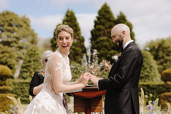 Ceremony moment at an outdoor wedding ceremony as groom places a ring on laughing bride in lace dress and veil beside a wooden podium in a garden