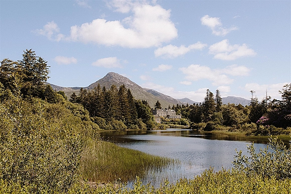 Mountain lake landscape with calm lake view reflecting distant peaks, framed by evergreens and reeds under a blue sky with clouds