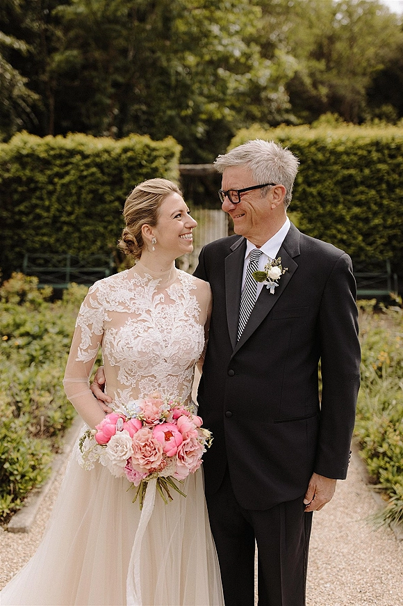 Wedding portrait of father and bride walking on a garden gravel path, her lace dress and pink bouquet beside his black suit and striped tie