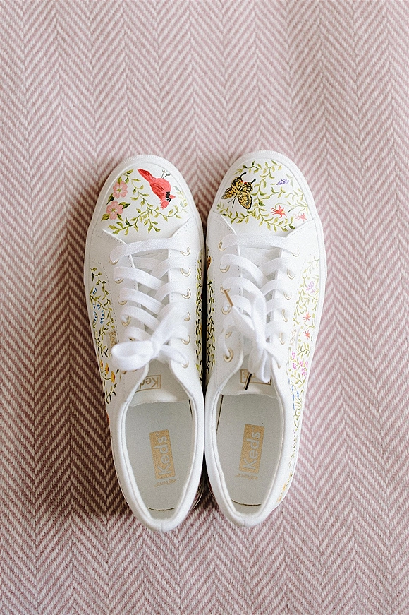 Bridal sneakers with floral print and bird and butterfly illustrations, white laces, laid on pink herringbone fabric background