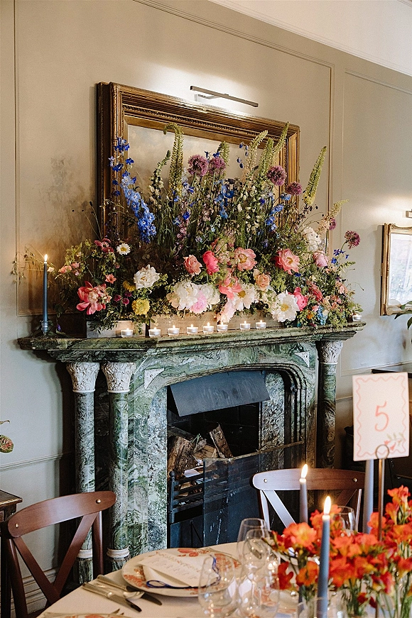 Wedding fireplace flowers with a mantel floral arrangement, taper and tealight candles, above a marble fireplace in a paneled dining room.