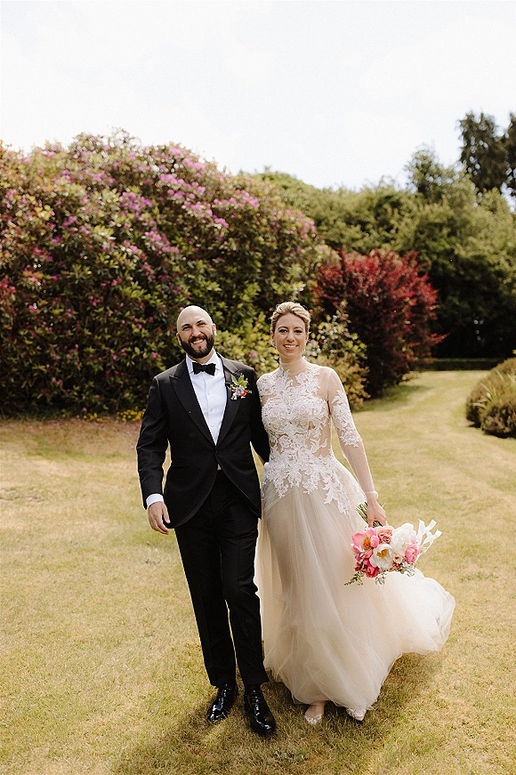 Couple portrait of bride and groom walking, her lace long-sleeve gown and pink-white bouquet, on a garden lawn with flowering bushes