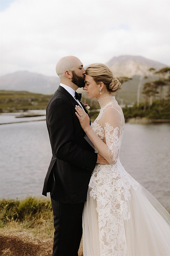 Couple portrait of groom kissing bride’s forehead as she rests her hand on his chest, lace sleeves visible by coastal mountains under clouds