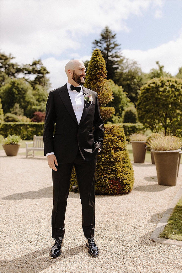 Groom portrait in a black tuxedo with bow tie and boutonniere, hands in pockets on a gravel garden path by topiary and bench