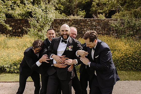 Groomsmen portrait of the wedding party laughing as they lift the groom in black tuxedos, in a garden by a stone wall