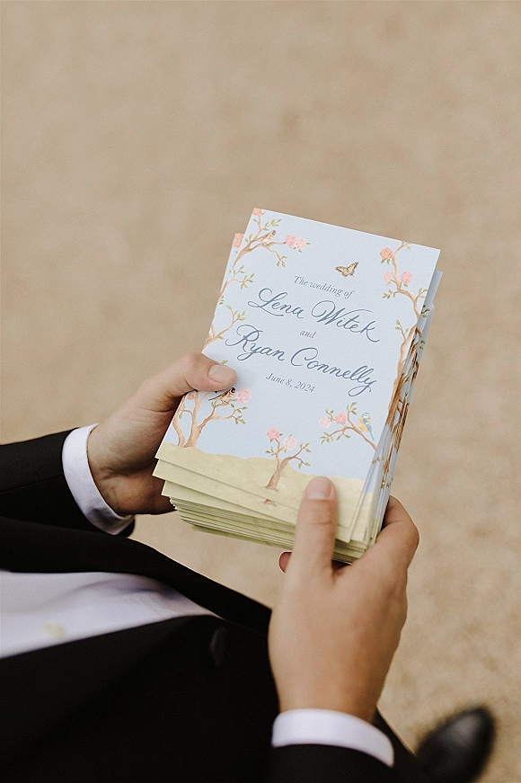 Wedding program with floral border and butterfly illustration in calligraphy, held over a neutral floor in soft natural light