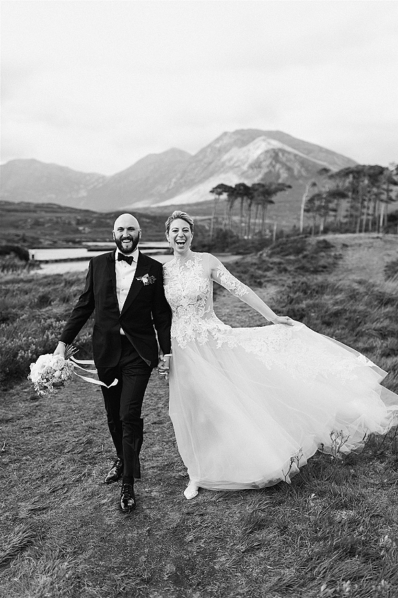 Couple portrait in a black and white wedding portrait style, bride in lace long sleeves holding bouquet, walking hand in hand by mountains and lake