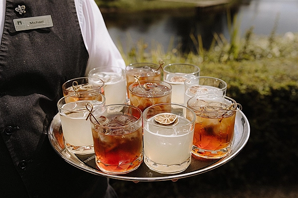 Wedding cocktails on a silver tray, signature wedding drinks in rocks glasses with citrus wheel garnish, carried by a vested bartender by greenery and water