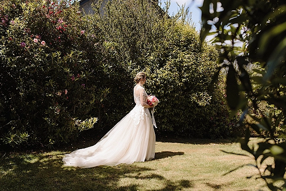 Bridal portrait of a bride holding bouquet, showing lace sleeves and long train in a sunlit garden with flowering shrubs and lawn
