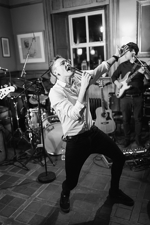 Wedding band performance with singer at a microphone and guitars beside a drum kit, playing in an indoor reception room with framed art