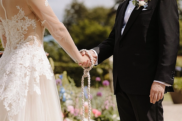 Wedding vows during a handfasting ceremony as bride in lace sleeves and groom in black tux hold hands bound with a braided cord outdoors