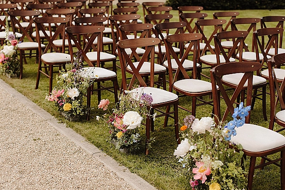Ceremony aisle decor with outdoor ceremony seating, cross back chairs and white cushions lined by low wildflower and garden rose florals on a gravel path