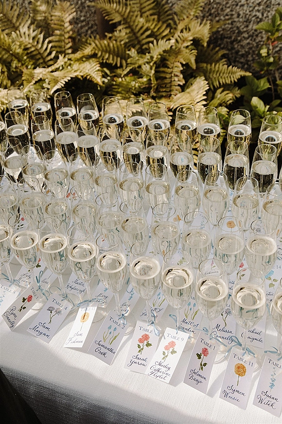 Wedding escort cards with calligraphy place cards tied in blue ribbon beside champagne flutes on a white tablecloth with fern greenery backdrop