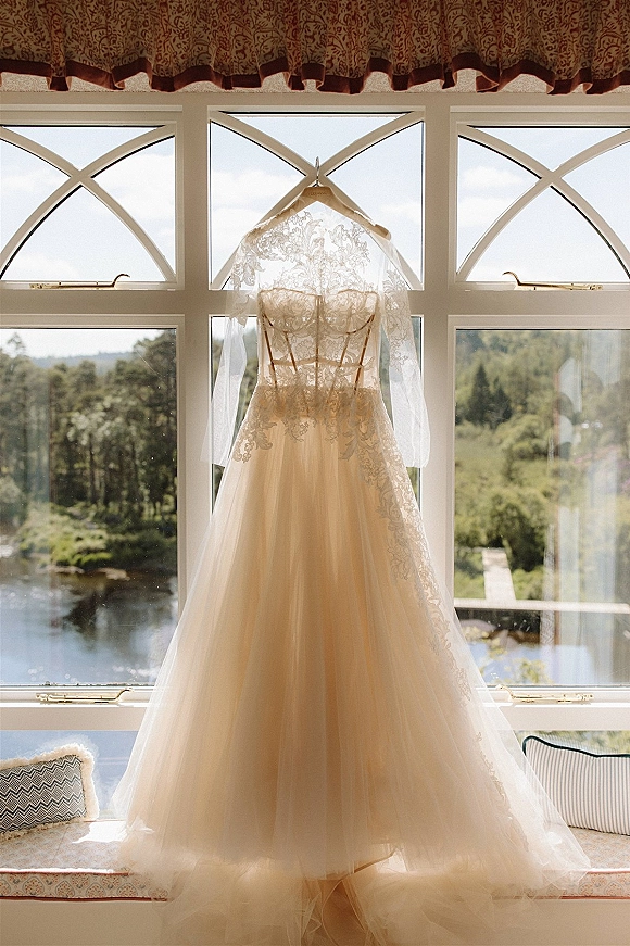 Wedding dress with lace wedding dress bodice and long sleeves on a hanger by large windows, tulle skirt framed by countryside view