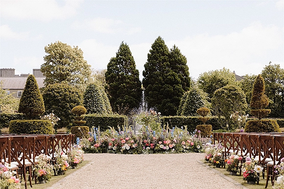 Ceremony setup with outdoor wedding ceremony aisle florals lining a gravel path, wooden chairs with white cushions in a formal garden by a fountain