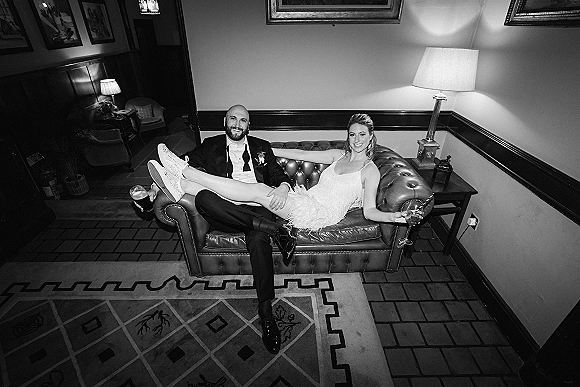 Couple portrait in a black and white wedding portrait style, bride and groom lounging on a leather sofa with wine and beer in hand.