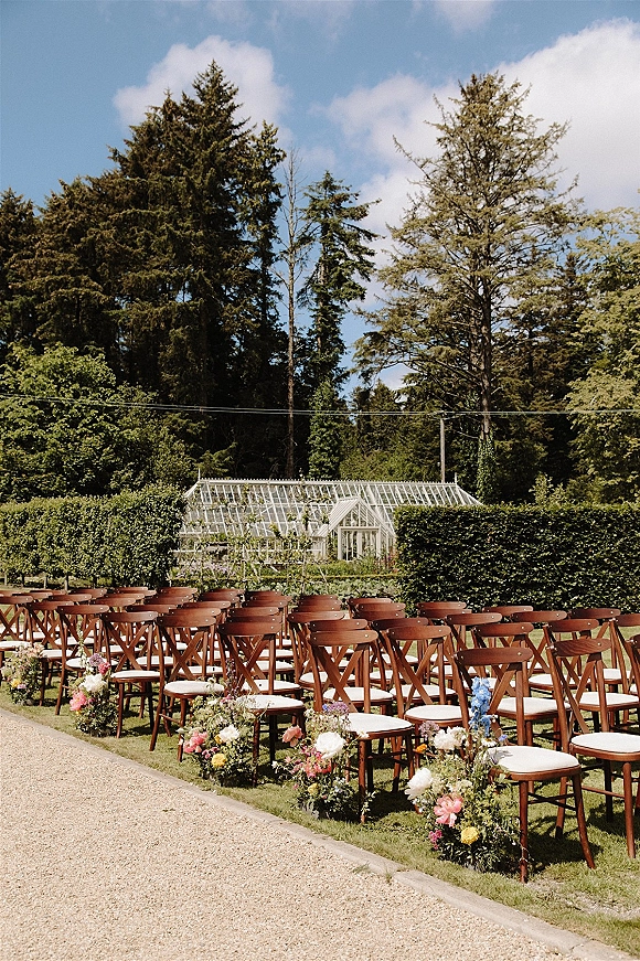 Ceremony seating with outdoor ceremony chairs in neat rows, white cushions and aisle flowers on a lawn beside a greenhouse and hedges