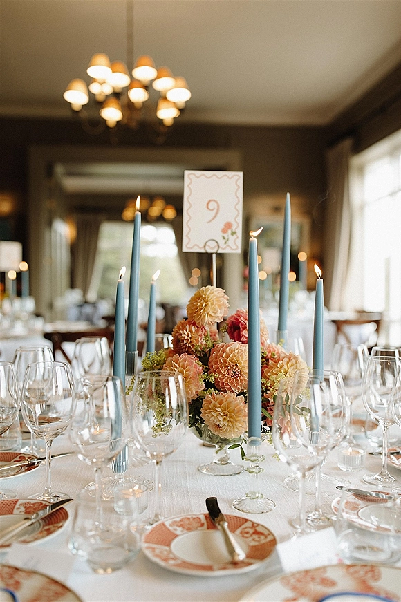 Reception tablescape with wedding table centerpiece, blue taper candles in glass holders, floral arrangement, patterned plates, and table number under chandeliers