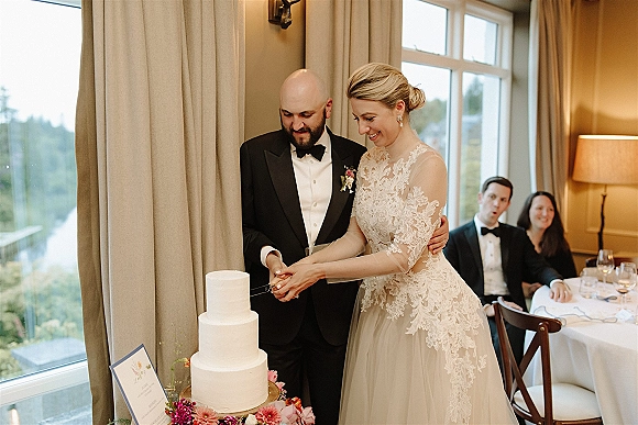 Cake cutting moment as bride and groom slice a two-tier wedding cake with fresh flowers in a window-lit reception room
