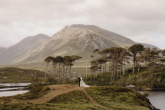 Couple portrait of bride and groom kissing beside a lake, her long veil and bouquet flowing on a grassy hillside under cloudy mountains