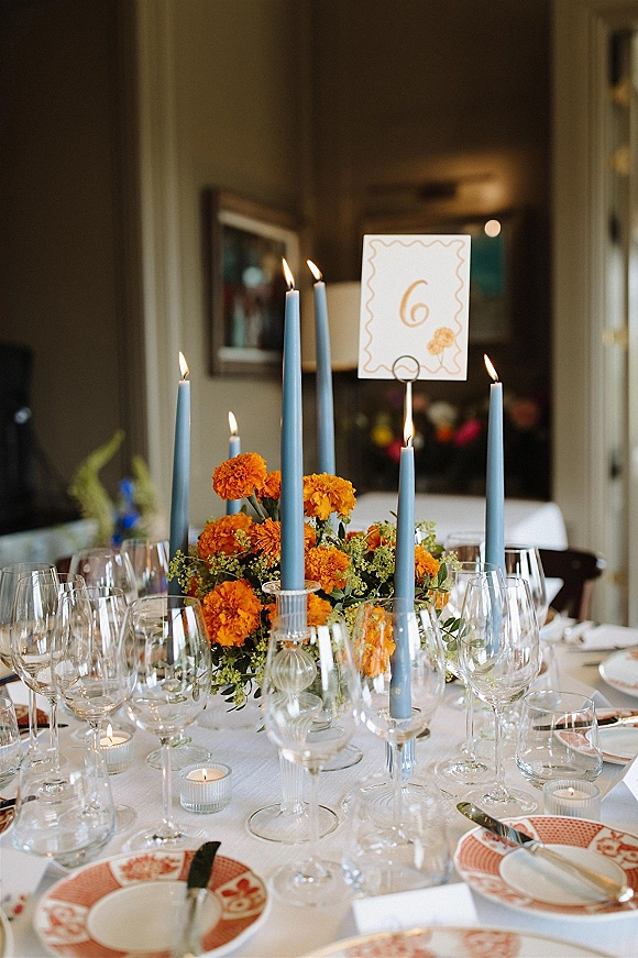 Reception tablescape with wedding table centerpiece of orange blooms, blue taper candles, patterned plates, and glassware in an indoor room