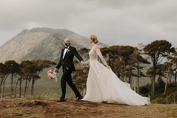 Couple portrait of bride and groom holding hands on a grassy mountainside, her lace gown with long train and bouquet under overcast sky