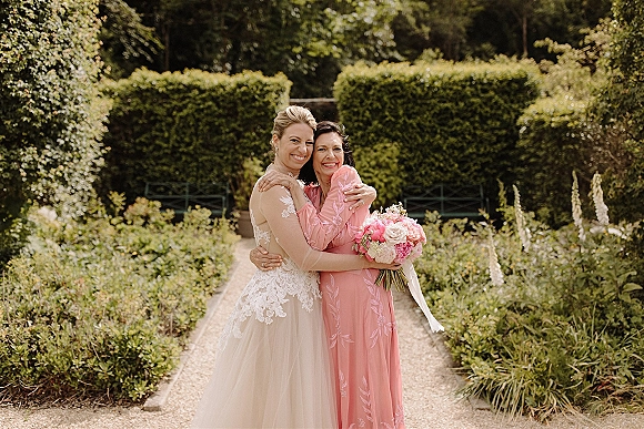 Bride with mother hugging on a garden path, holding a pink and white bouquet with ribbon wrap, surrounded by hedges and greenery