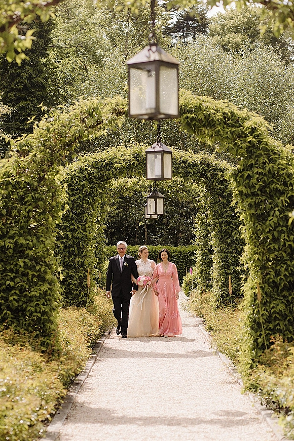 Processional with bride walking down aisle, holding a bouquet with ribbon, escorted on a garden path beneath a hedge archway of lanterns