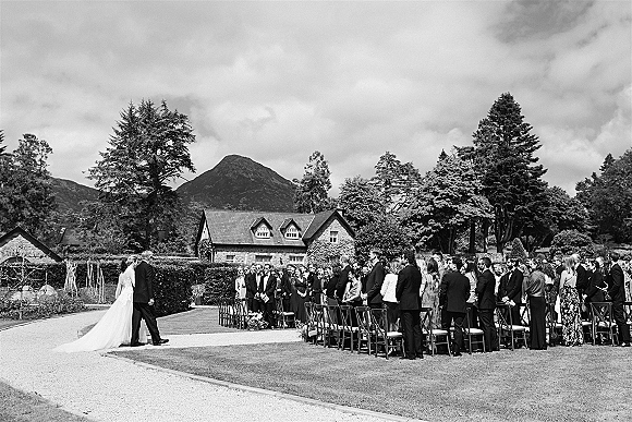 Wedding processional as the bride walking down aisle in a veil and long train on a gravel path past guests, with mountains beyond