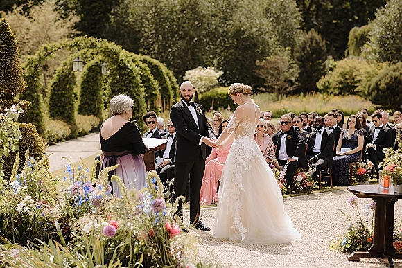 Wedding vows as bride in lace dress and groom in tuxedo hold hands while officiant reads at a garden ceremony with guests and aisle florals
