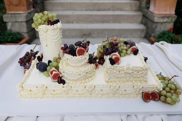 Wedding cake, fruit-topped and edged with buttercream piping, set on a white tablecloth by stone steps in a leafy garden.