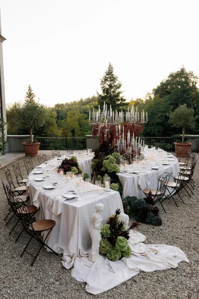 Reception tablescape for an outdoor reception table, crystal candelabras casting warm light in a gravel courtyard framed by potted trees at dusk.
