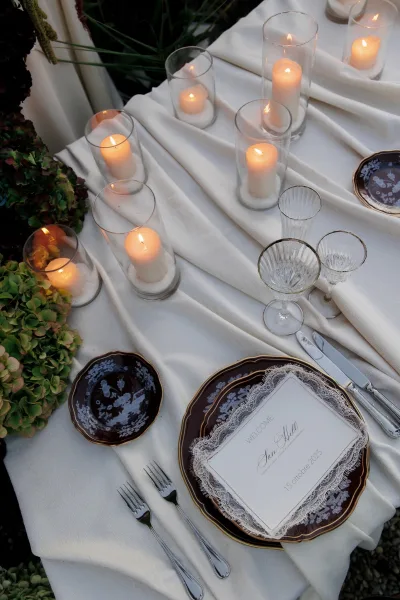 Wedding place setting with layered plates, softly lit by pillar candles, set amid garden greenery on gravel ground.