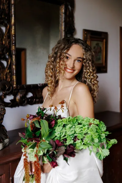 Bridal portrait of a bride holding bouquet, with floral embroidery on her bodice, framed by an ornate mirror and wooden dresser in a white room.