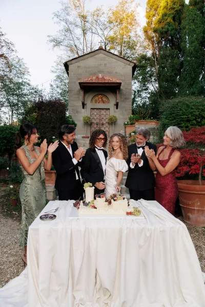 Wedding cake cutting as the bride holds the cake knife at a simple cake table set beside a stone chapel amid garden trees and potted plants.