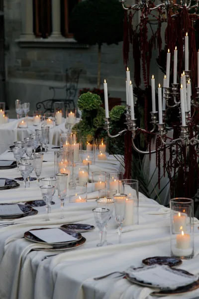 Reception tablescape glowing as a candlelit reception table, anchored by a candelabra, in a courtyard with a stone wall, topiary, and greenery.