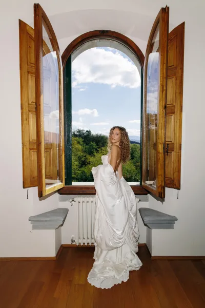 Bridal portrait of a bride by window, framed by an arched window overlooking hills, wearing a low-back gown.