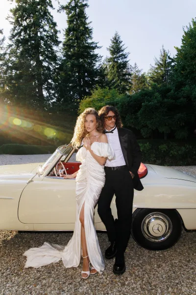 Couple portrait of a bride and groom with classic car, beside the classic convertible on a gravel driveway framed by evergreen trees.