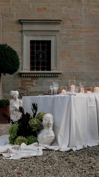 Reception tablescape with candlelit table decor centered on a bust sculpture, set in a gravel courtyard against a stone wall.