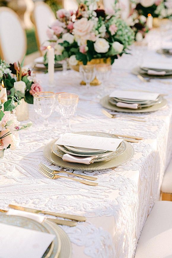 Reception tablescape with lace wedding tablecloth, pink and white centerpiece, taper candles, gold flatware, and patterned plates on an outdoor lawn
