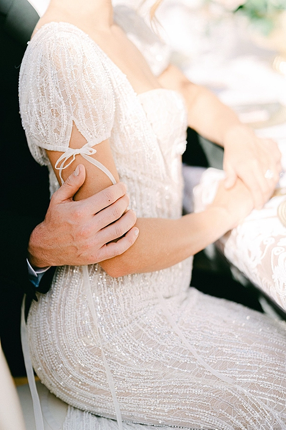 Couple portrait with wedding couple close up as the groom holds the bride’s hand, highlighting her beaded lace dress and ring at a reception table