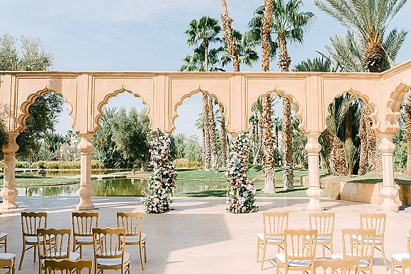 Ceremony setup with floral pillars and rows of chairs facing stone arches, beside a reflecting pool under palm trees and blue sky